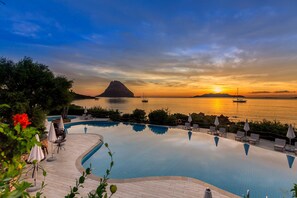 A swimming pool with lounge chairs and umbrellas, surrounded by greenery, with a sunset view over water and a distant mountain. - Hotel Don Diego (Loiri Porto San Paolo)