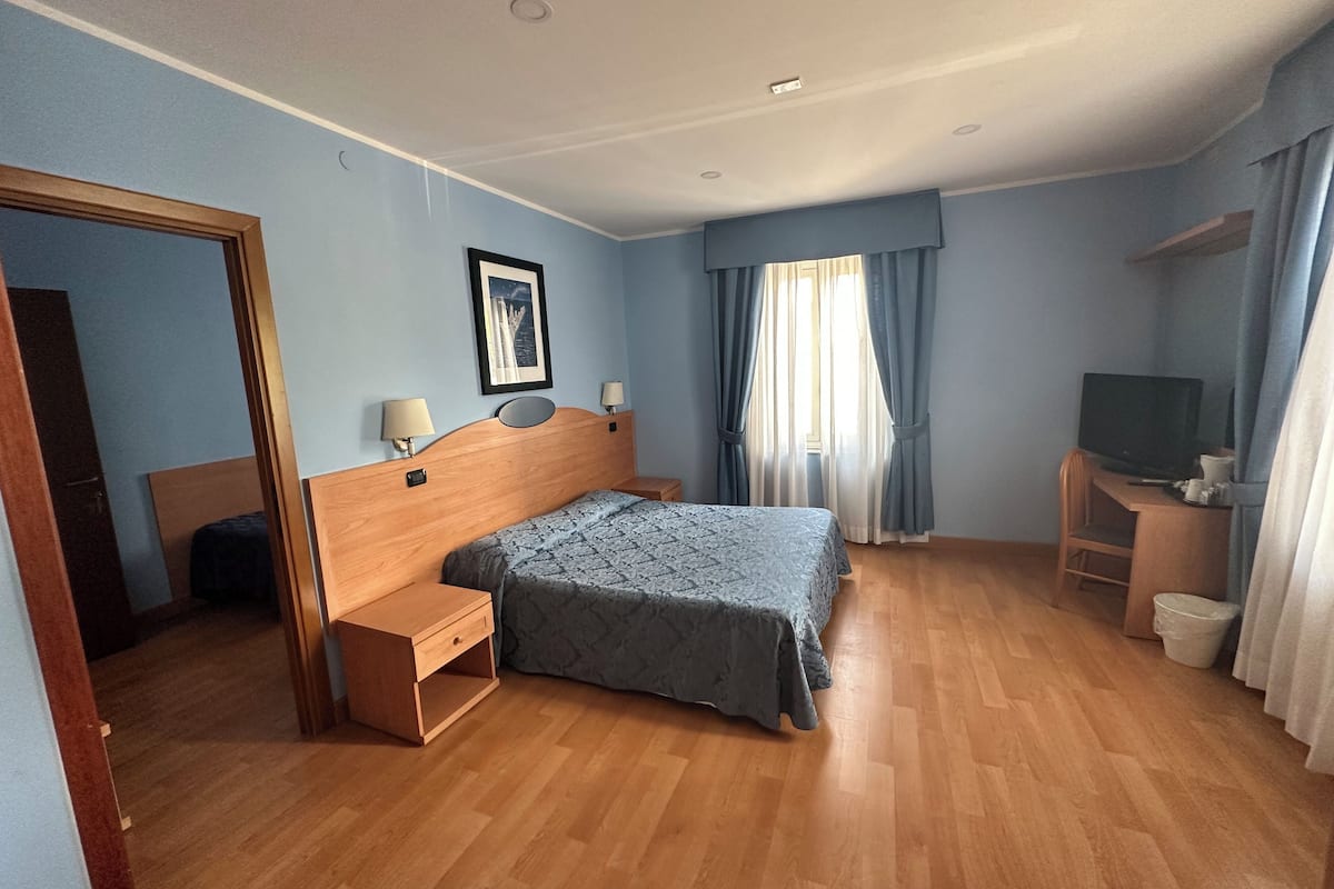 A hotel room with a bed, bedside tables, a desk with a chair, a television, and a window with curtains.