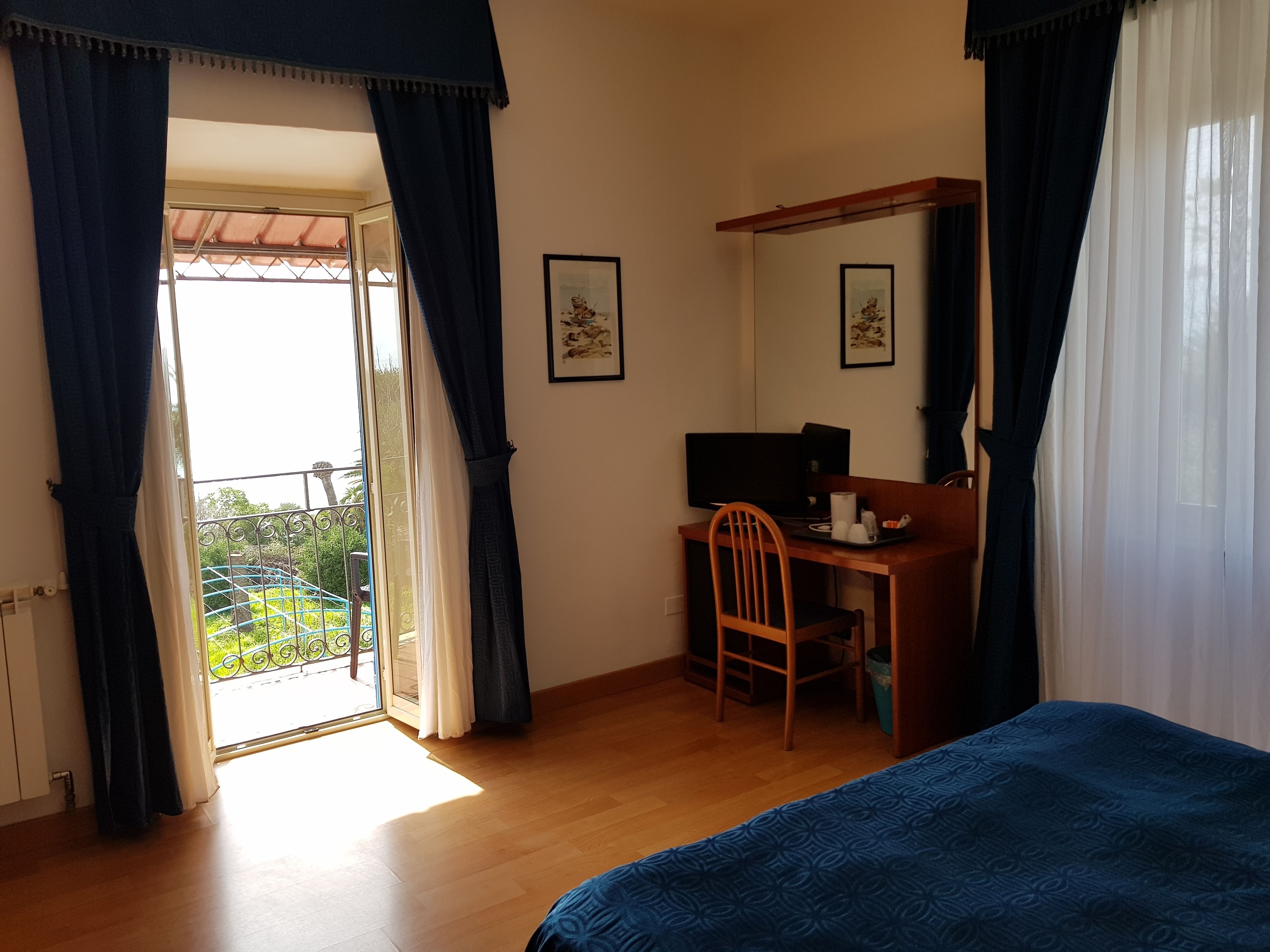 Habitación de hotel con cama, escritorio con silla, televisor y balcón con cortinas.
