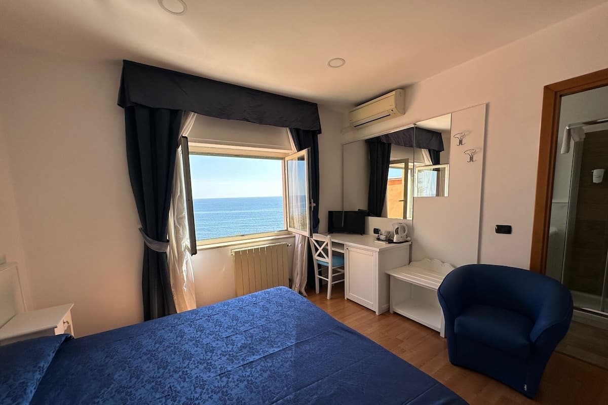 A bedroom with a bed, a desk, a chair, and a view of the sea.