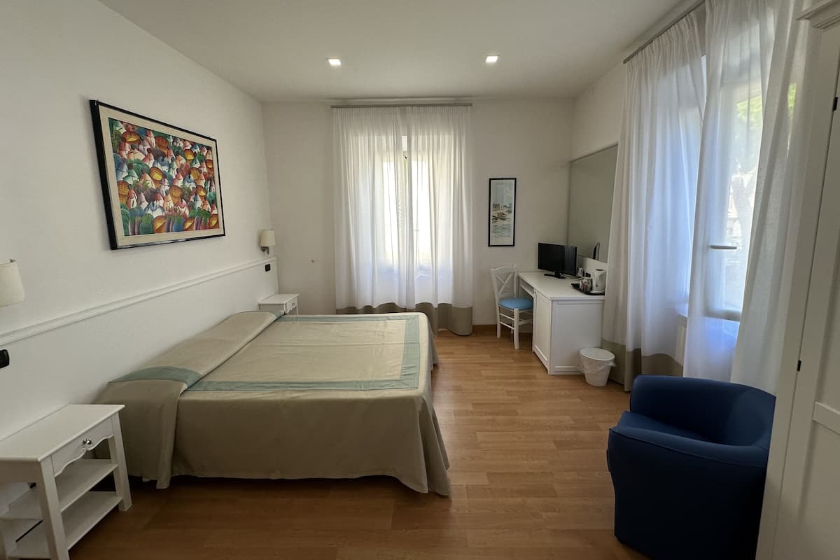 A hotel room with a bed, a desk, a chair, a TV, and a painting on the wall.