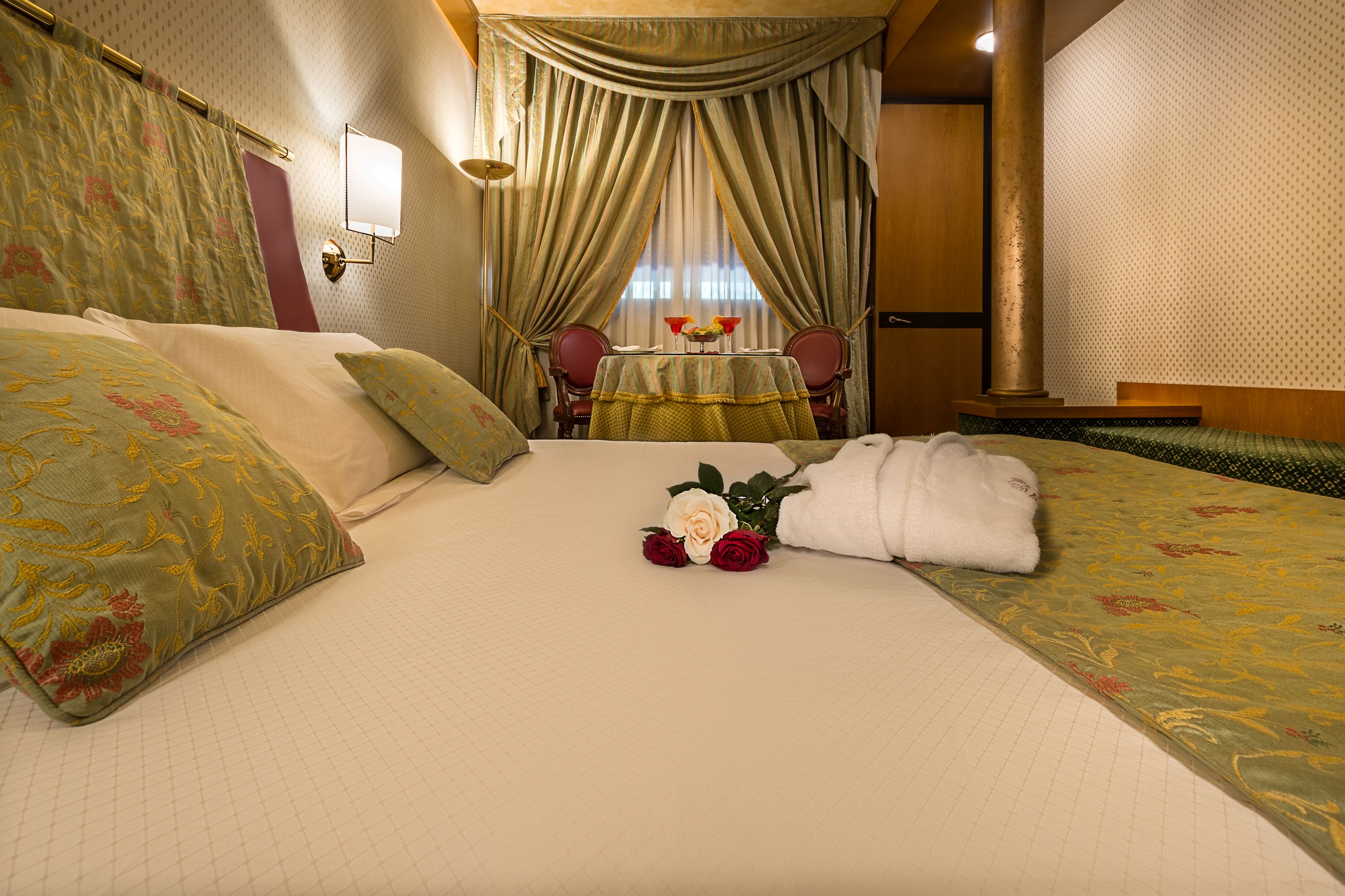 A hotel room with a large bed, floral bedspread, a table with a vase of flowers, and a view of the city through the window.