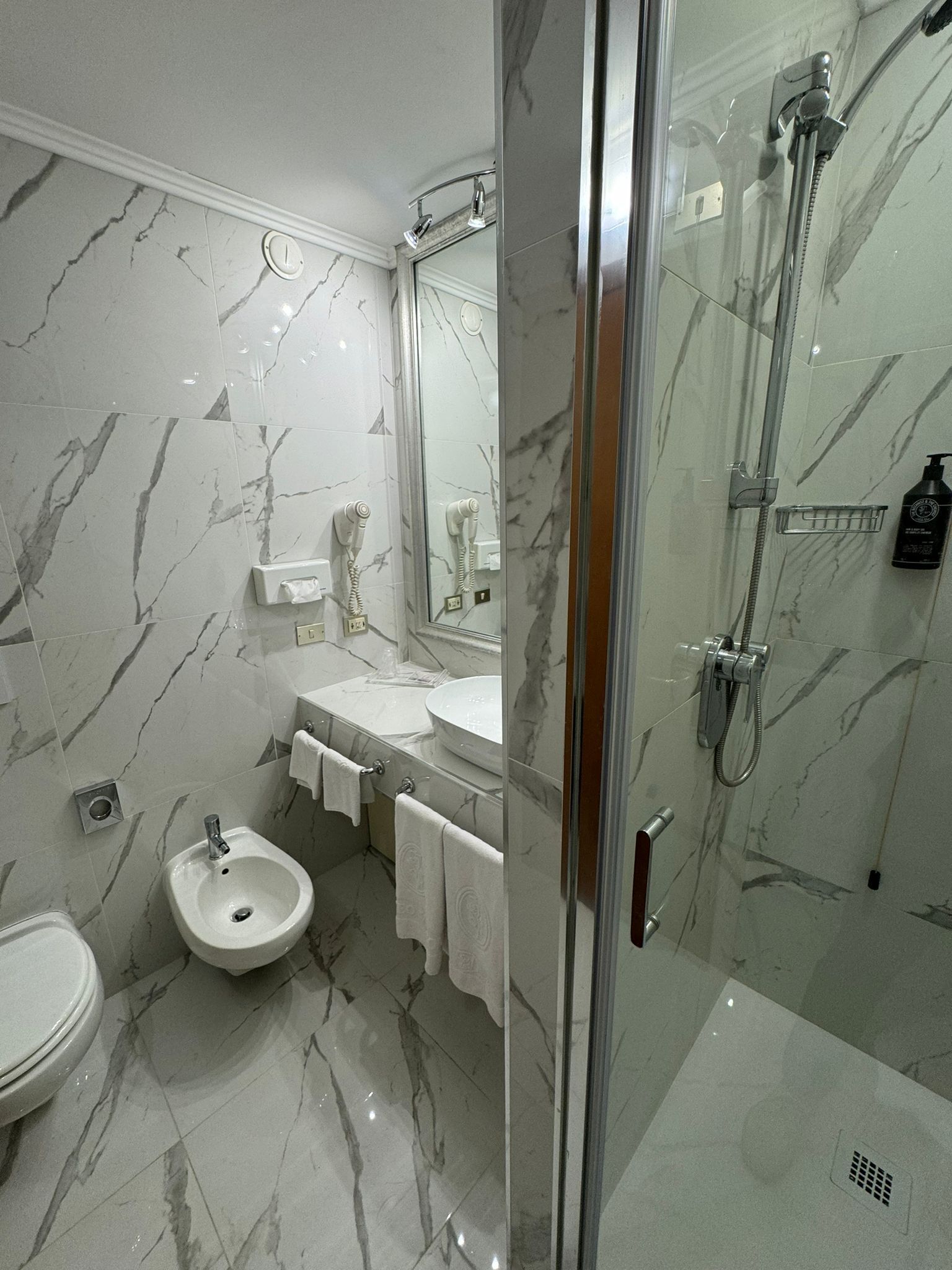 Un baño moderno con suelo de mármol, ducha con cerramiento de vidrio, un bidé y un lavamanos con espejo.