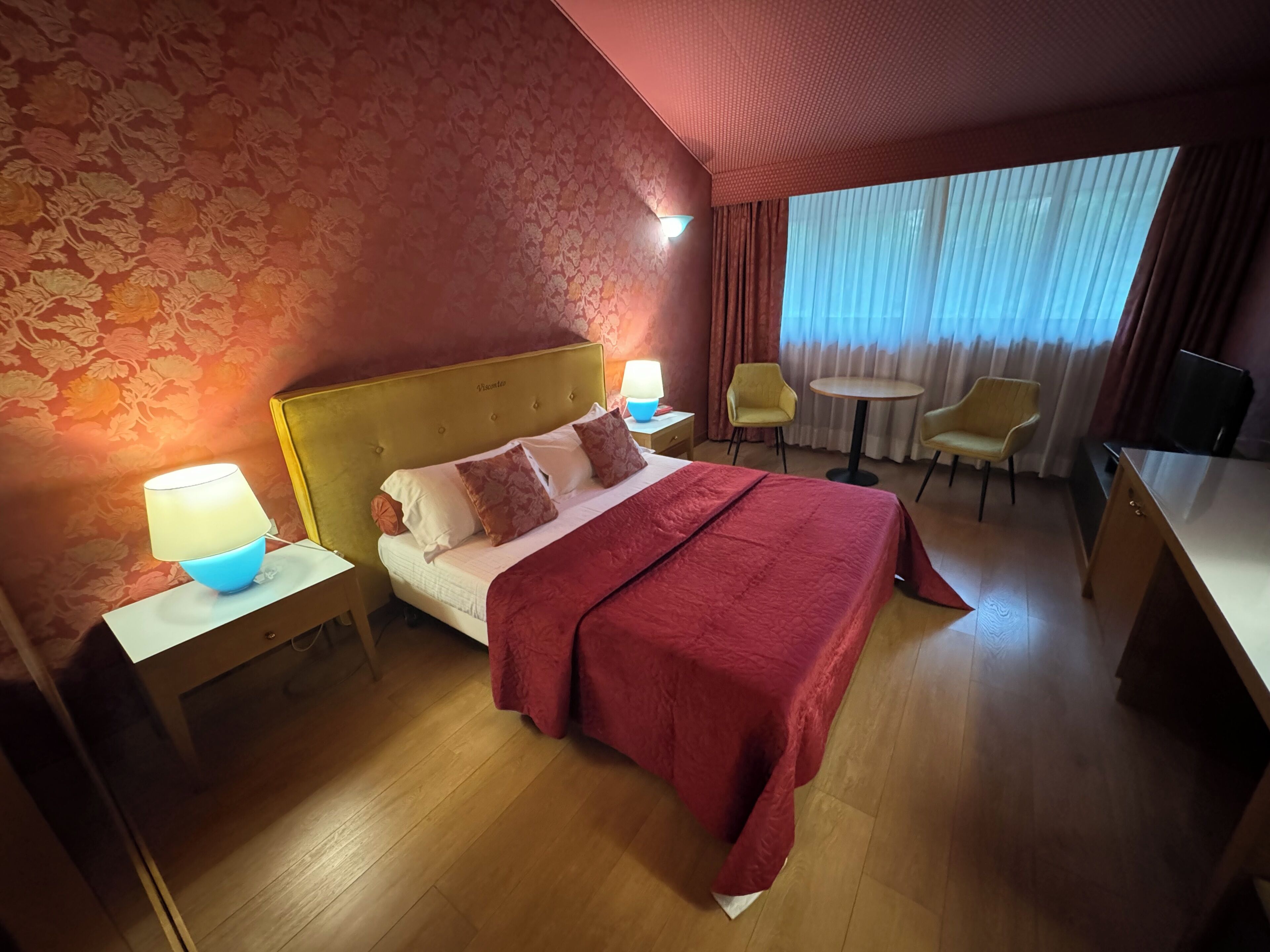 Habitación de hotel con cama, mesitas de noche, lámpara, silla, pequeña mesa y televisor.