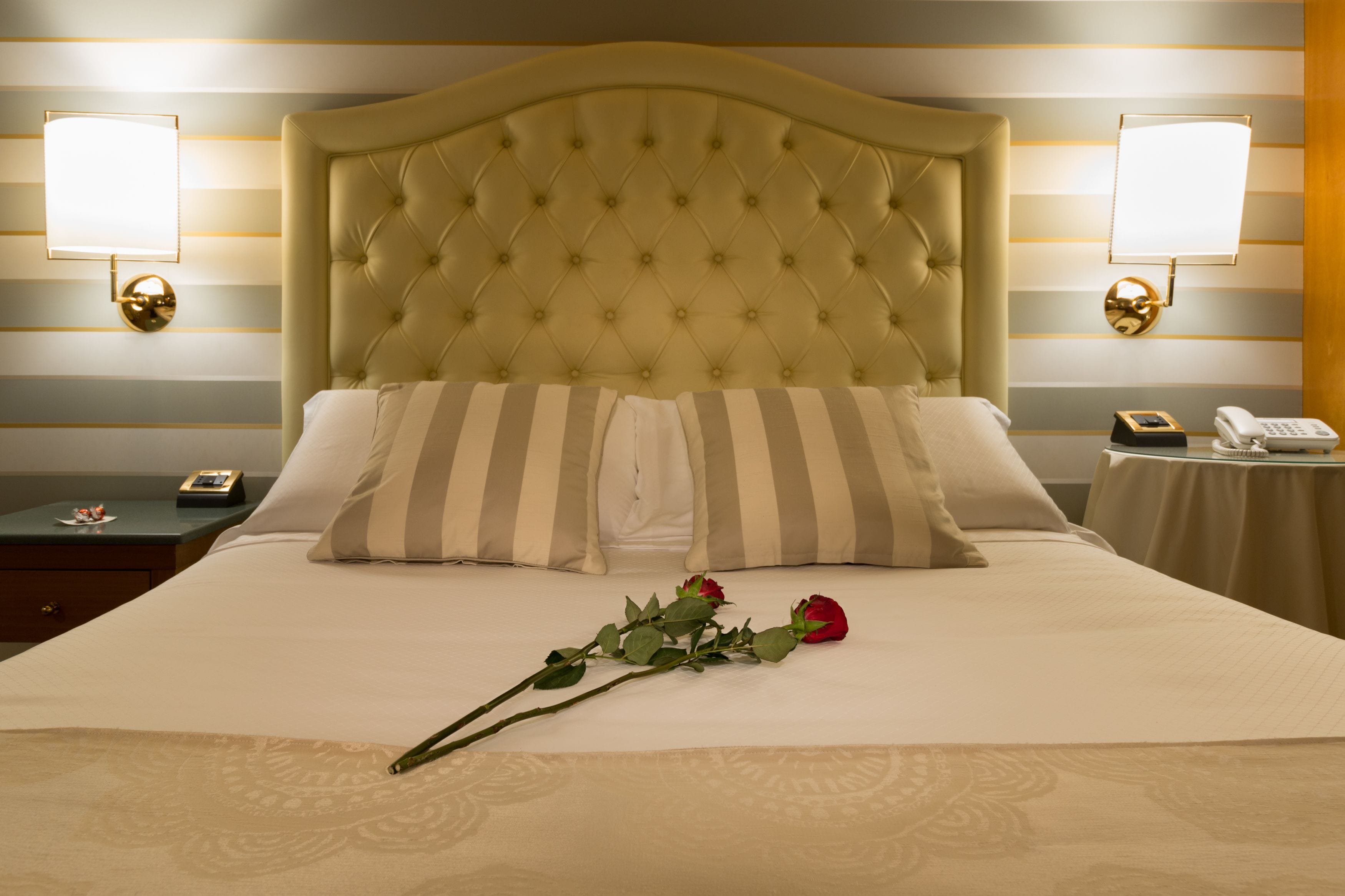 A hotel room with a tufted headboard, striped pillows, a bedside table with a lamp, a telephone, and a rose on the bed.