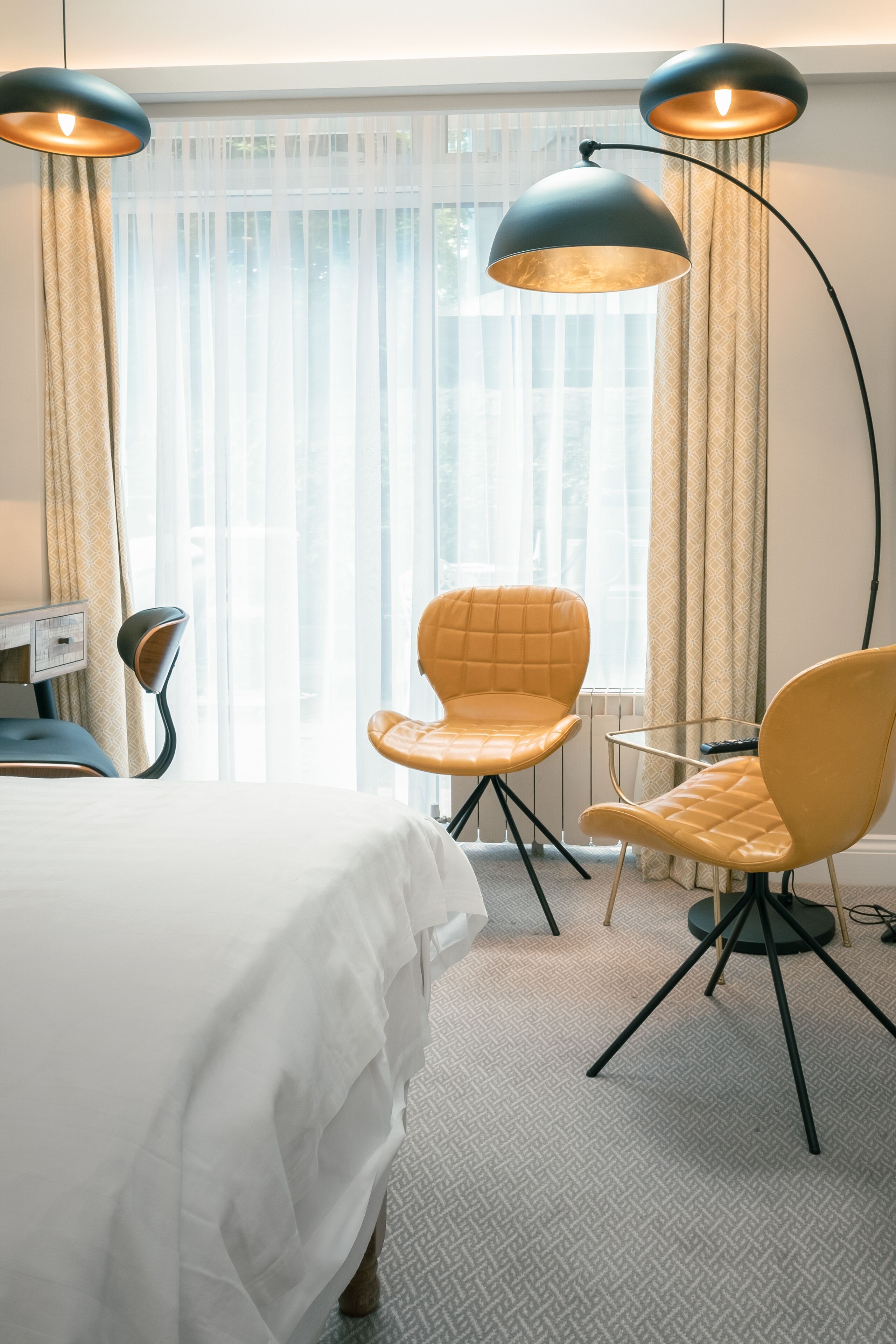 A hotel room with a bed, two yellow chairs, a desk, and two pendant lights.