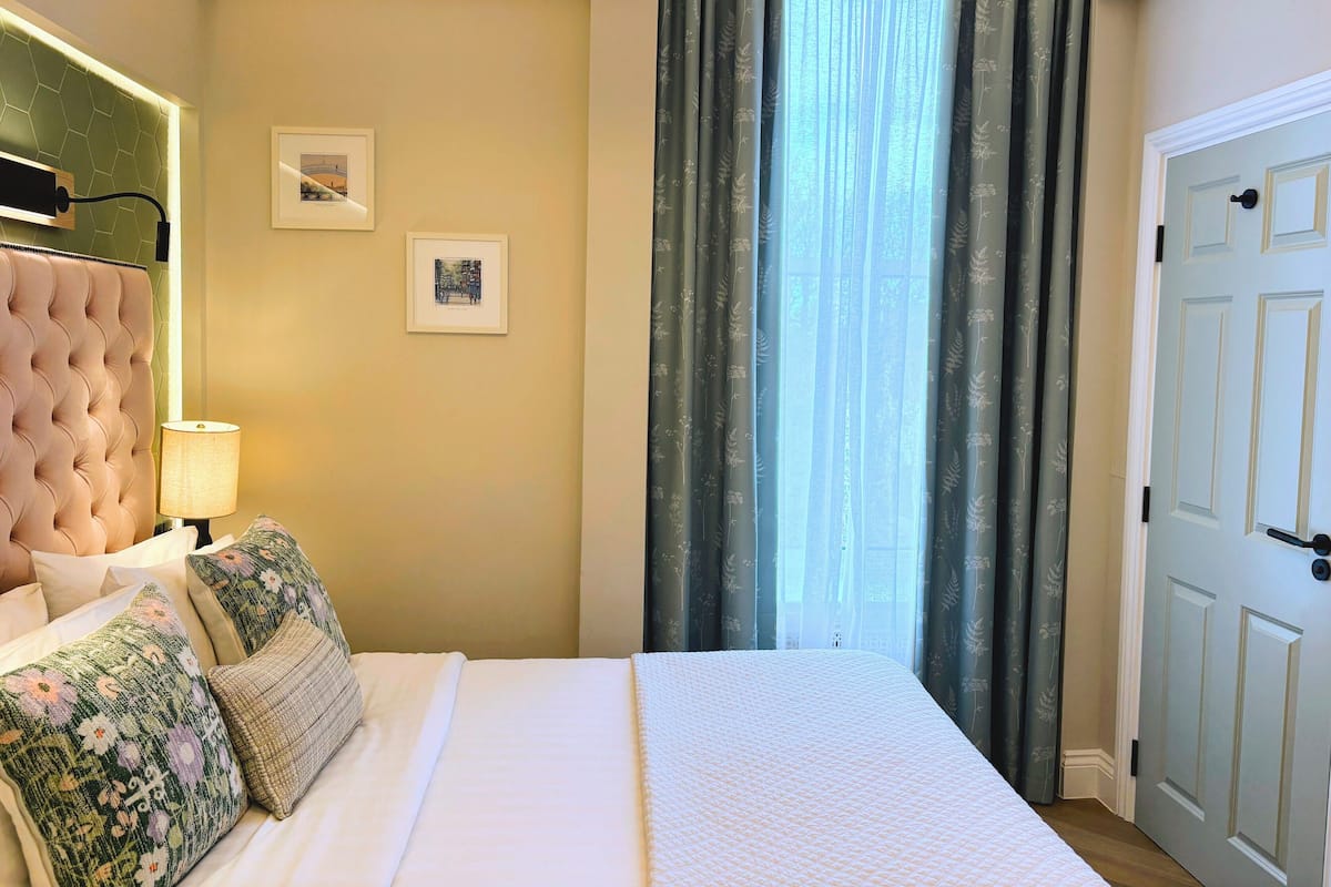 Quarto com cabeceira estofada, cama com roupa de cama branca, almofadas com padrão floral, abajur e duas fotos emolduradas na parede.