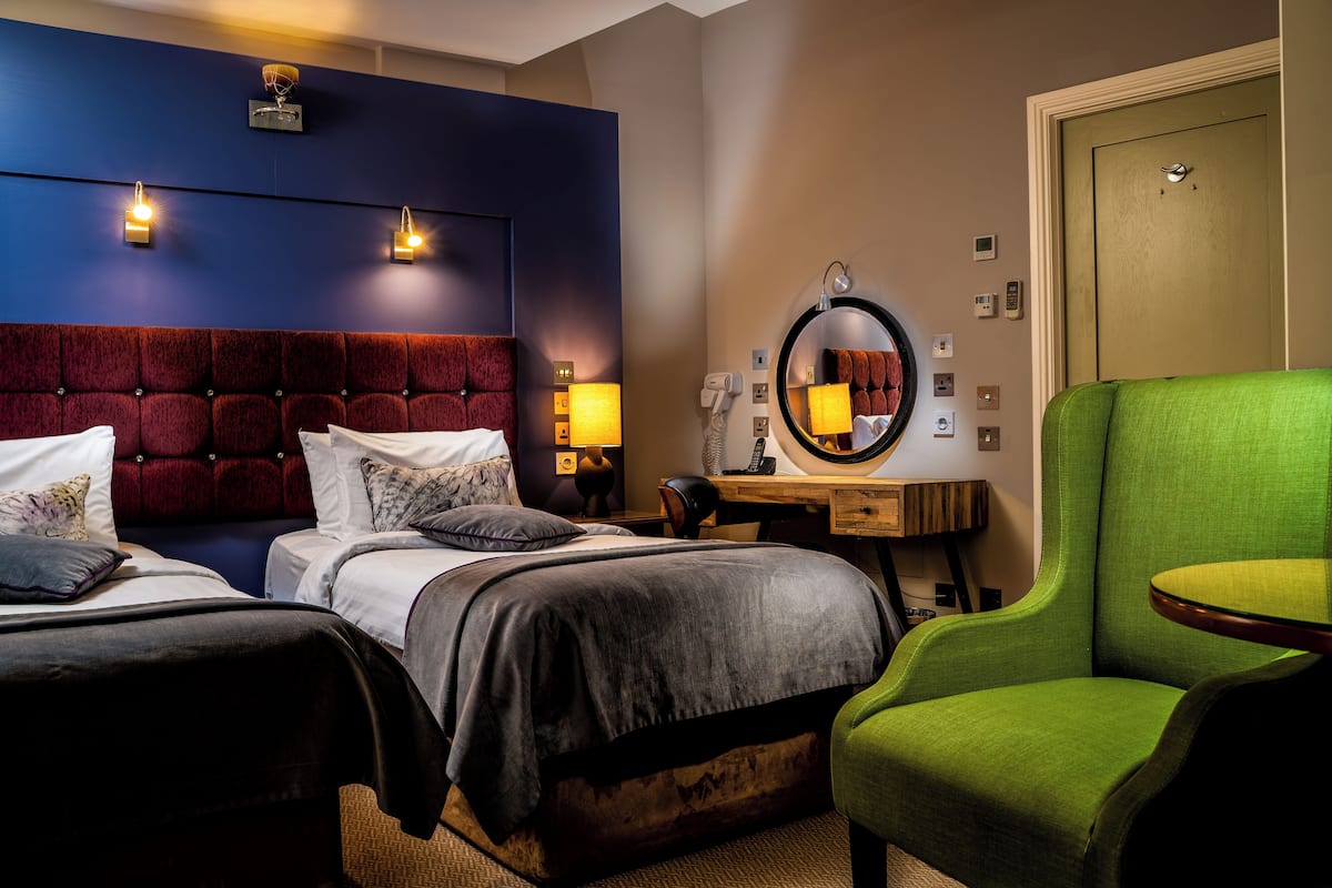 Quarto de hotel com duas camas, uma poltrona verde, uma escrivaninha com espelho e uma porta.
