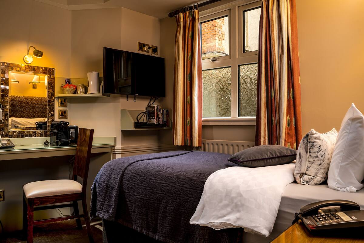 Quarto de hotel com cama, escrivaninha, cadeira, televisão e janela com cortinas.