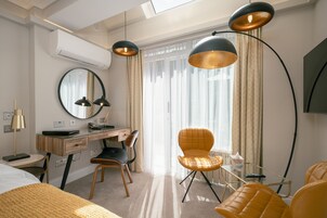 Quarto de hotel moderno com escrivaninha, duas cadeiras, uma cama e uma janela grande com cortinas.