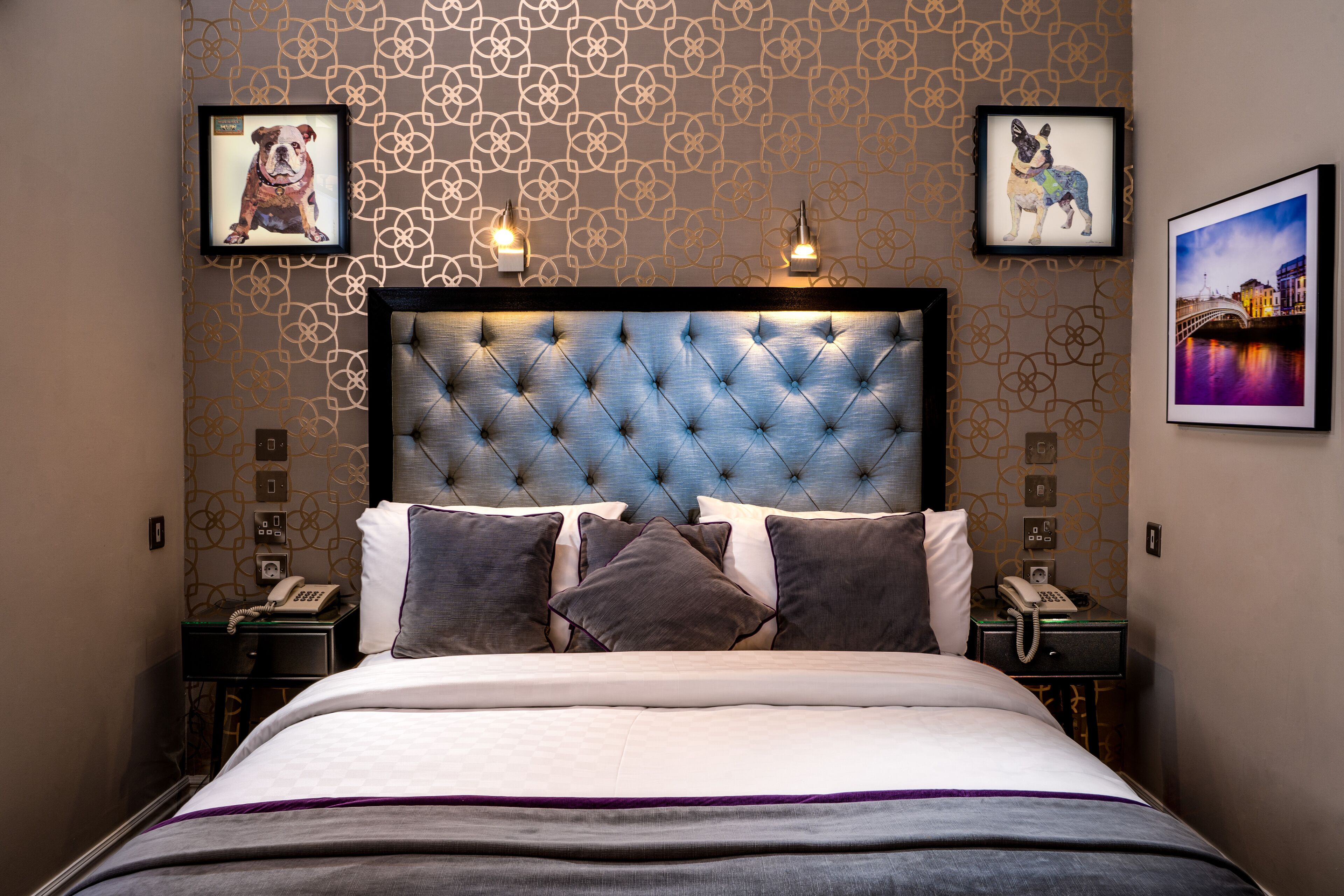 Habitación de hotel con cabecera capitonada, cama con colcha blanca y morada, dos mesitas de noche con lámparas y cuadros enmarcados en las paredes.