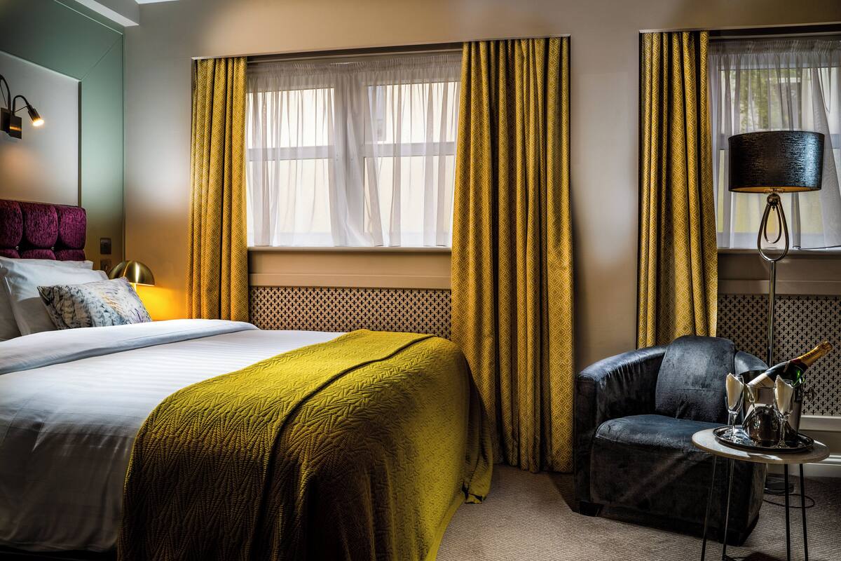 Quarto de hotel com cama, cadeira, luminária e janela com cortinas.