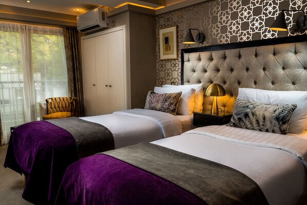A hotel room with two beds, a large window with curtains, a wall-mounted air conditioner, and a decorative headboard.