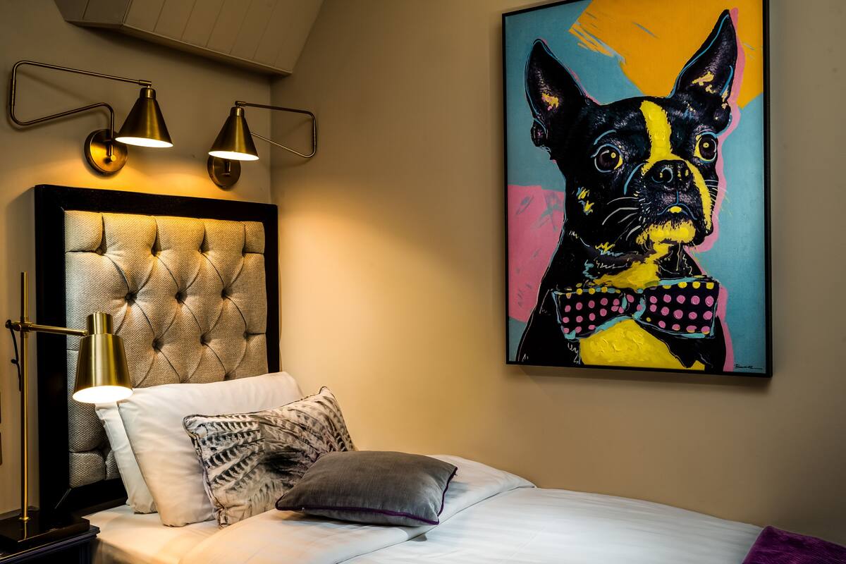 Quarto com cabeceira estofada, cama com roupa de cama branca, travesseiro cinza e pintura pop art de um cachorro emoldurada.