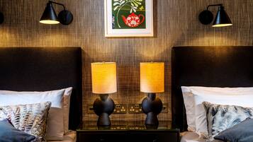 A hotel room with two beds, each with a headboard, a bedside table, and two lamps. There is a framed artwork above the beds.
