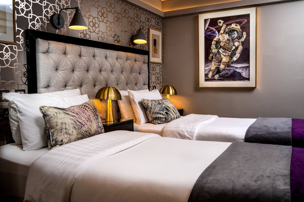 Quarto de hotel com cabeceira estofada, duas mesas de cabeceira com luminárias, uma obra de arte com tema de astronauta emoldurada, e cama com roupa de cama branca e manta roxa.