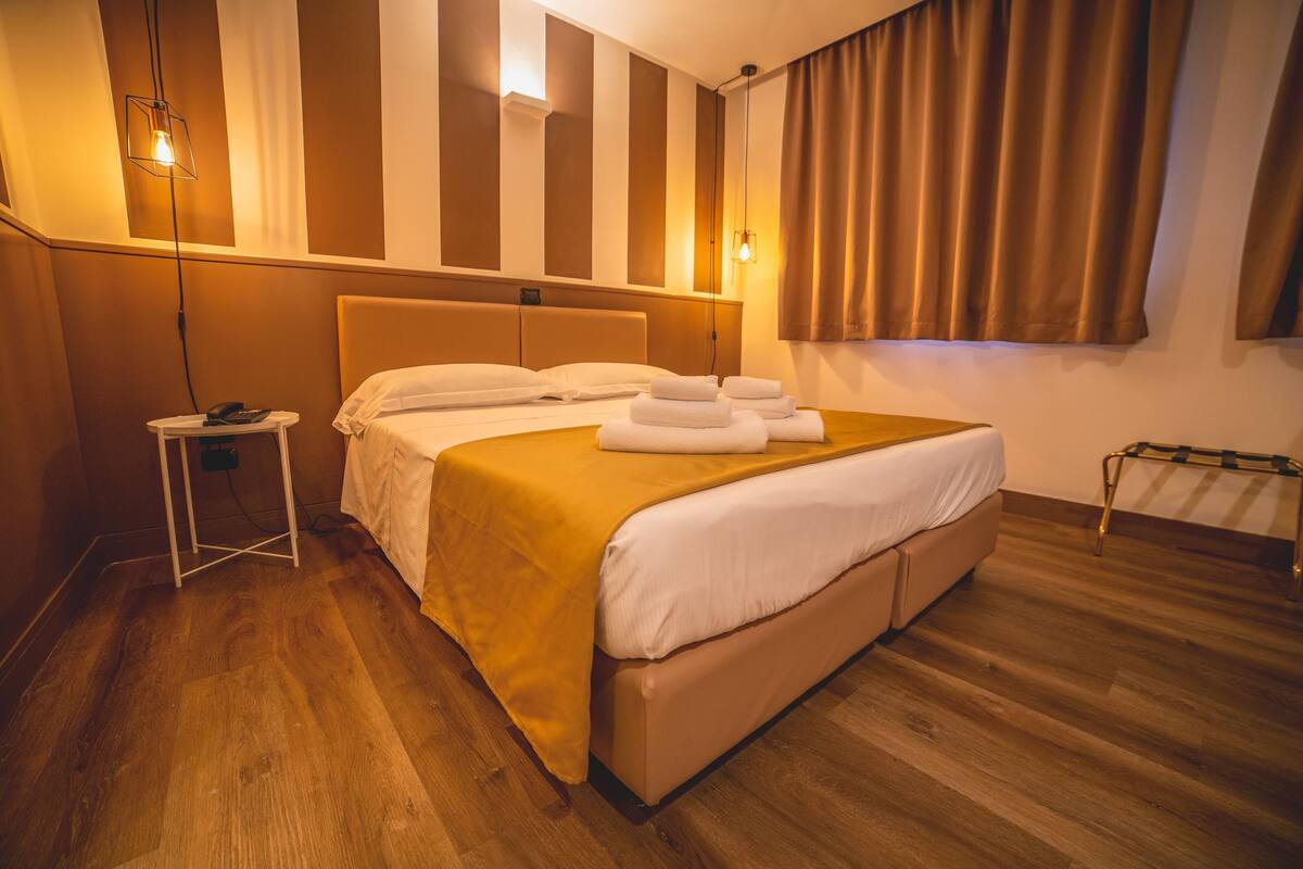 Een hotelkamer met een groot bed, een nachtkastje, een lamp en een raam met gordijnen.