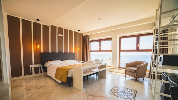A modern hotel room with a large bed, a seating area, a view of the sea, and a balcony.