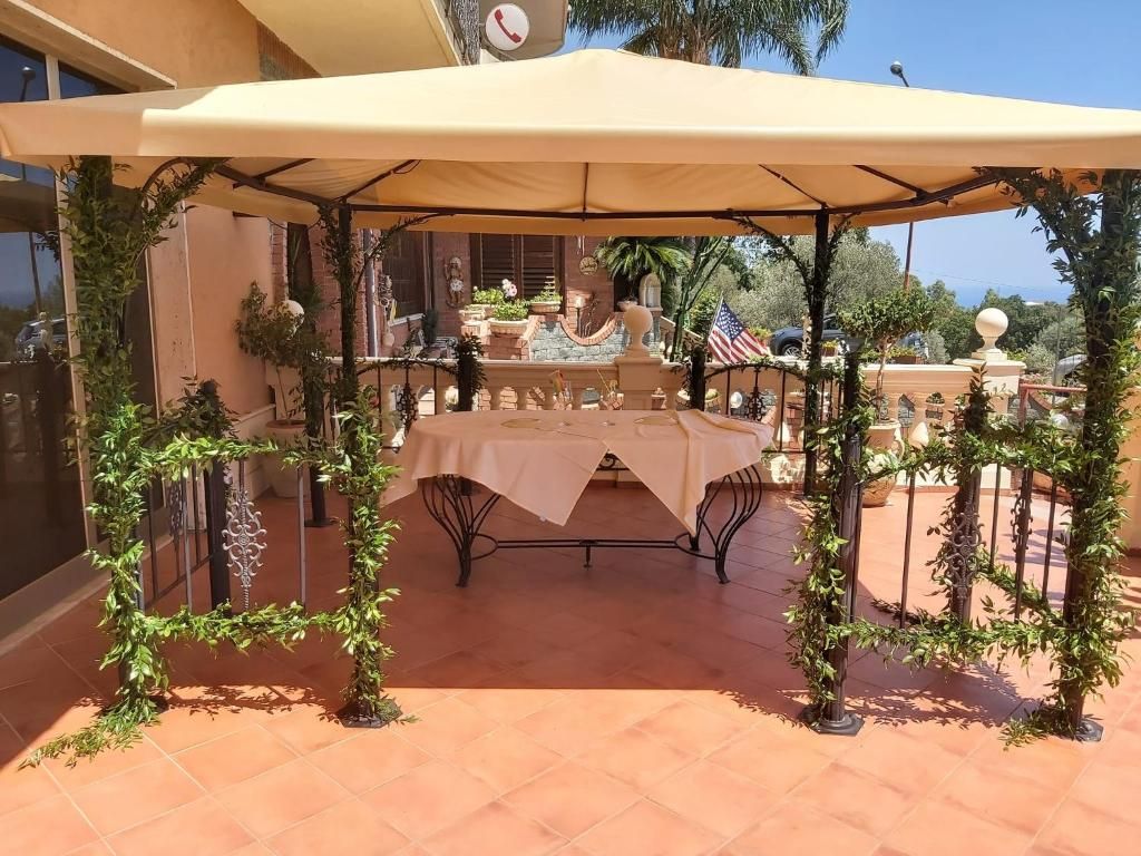 Pátio externo com mesa e cadeiras sob um toldo, cercado por vegetação e um cercado decorativo.
