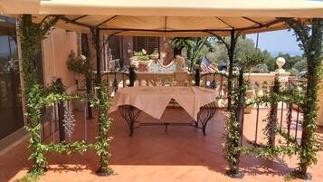 Pátio externo com mesa e cadeiras sob um toldo, cercado por vegetação e um cercado decorativo.
