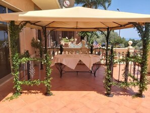 An outdoor patio with a table and chairs under a canopy, surrounded by greenery and a decorative fence. - Casa del Gourmet (Siderno)