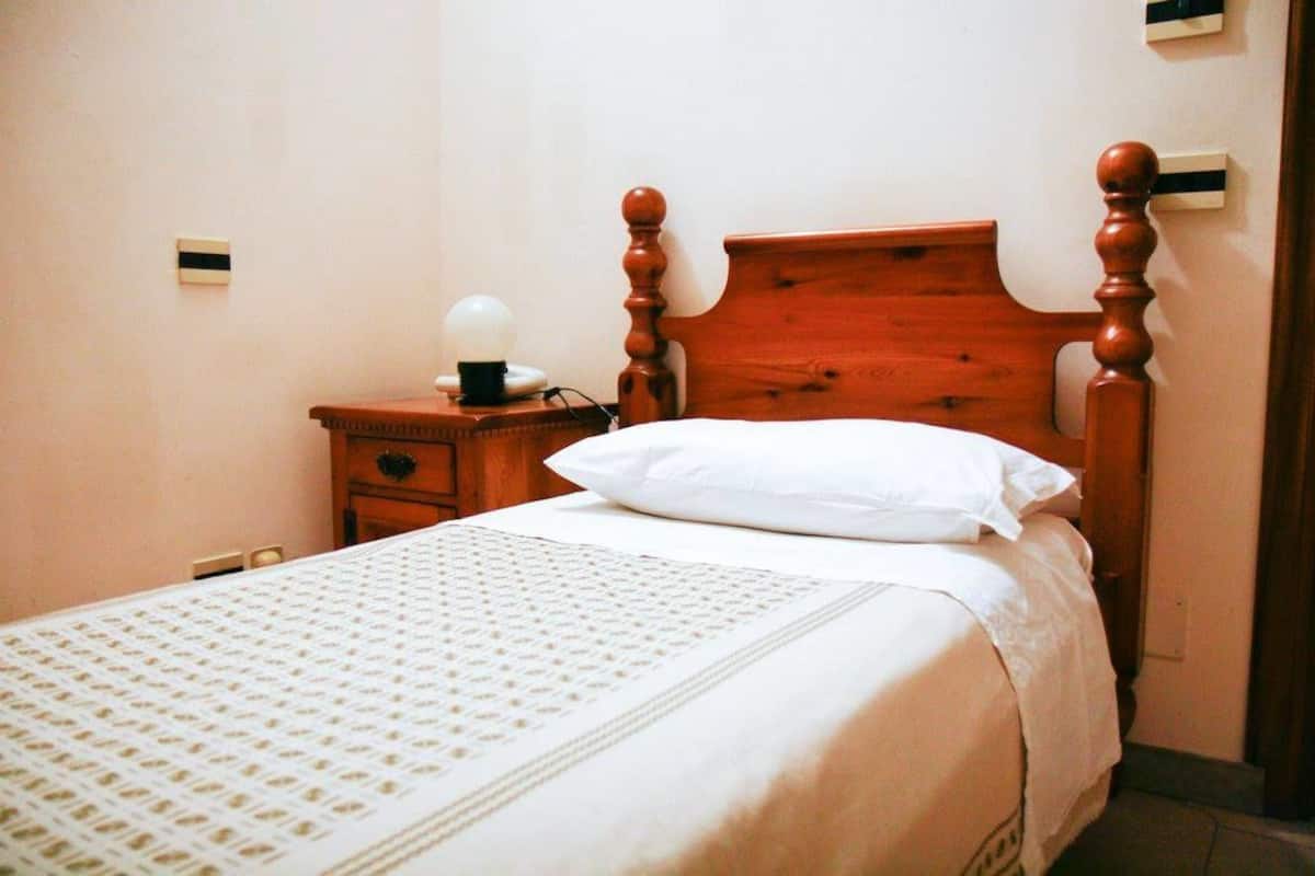 A neatly made bed with a wooden headboard, a bedside table with a lamp, and a wall-mounted light switch.
