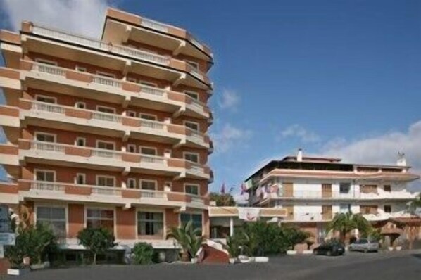 Un hotel de varios pisos con balcones, un estacionamiento y algo de vegetación.