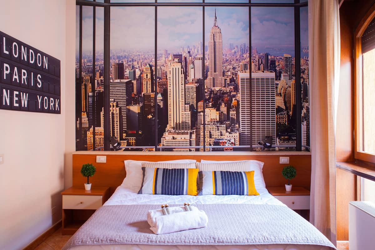 A hotel room with a large cityscape view, a bed with white bedding, two bedside tables with plants, and a sign listing London, Paris, and New York.