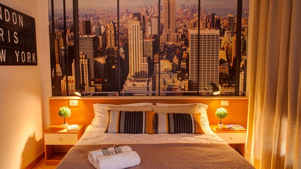A hotel room with a large cityscape view, a neatly made bed, two bedside tables with lamps, and a sign on the wall. - Hotel Ares (Milan)