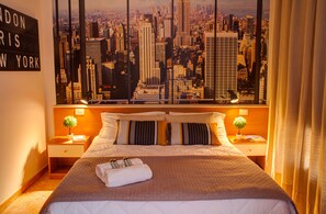 Habitación de hotel con amplia vista panorámica de la ciudad, cama bien hecha, dos mesitas de noche con lámparas y un cartel en la pared.