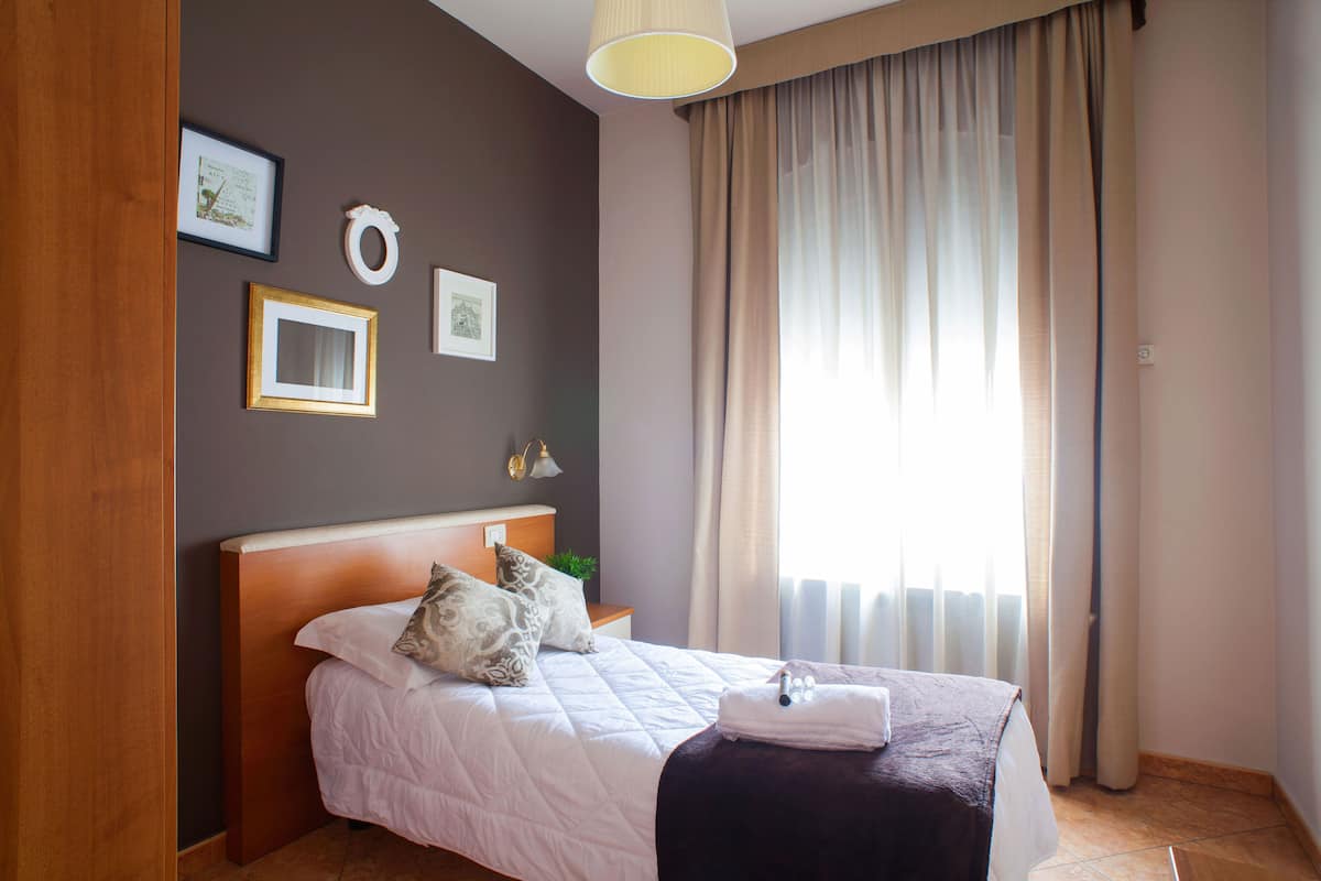 A neatly made bed with a white duvet, a dark throw blanket, and a set of towels. There are two framed pictures on the wall, a small plant on the nightstand, and a lamp.