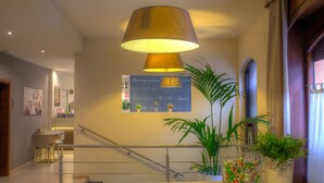 A hotel lobby with a staircase, a large hanging lamp, a signboard with amenities, and potted plants. - Hotel Ares (Milan)