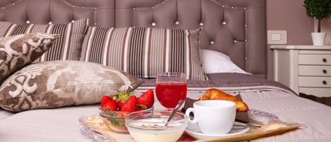 A neatly made bed with striped pillows, a bedside table with a lamp, and a tray with breakfast items including strawberries, a glass of red juice, and a croissant.