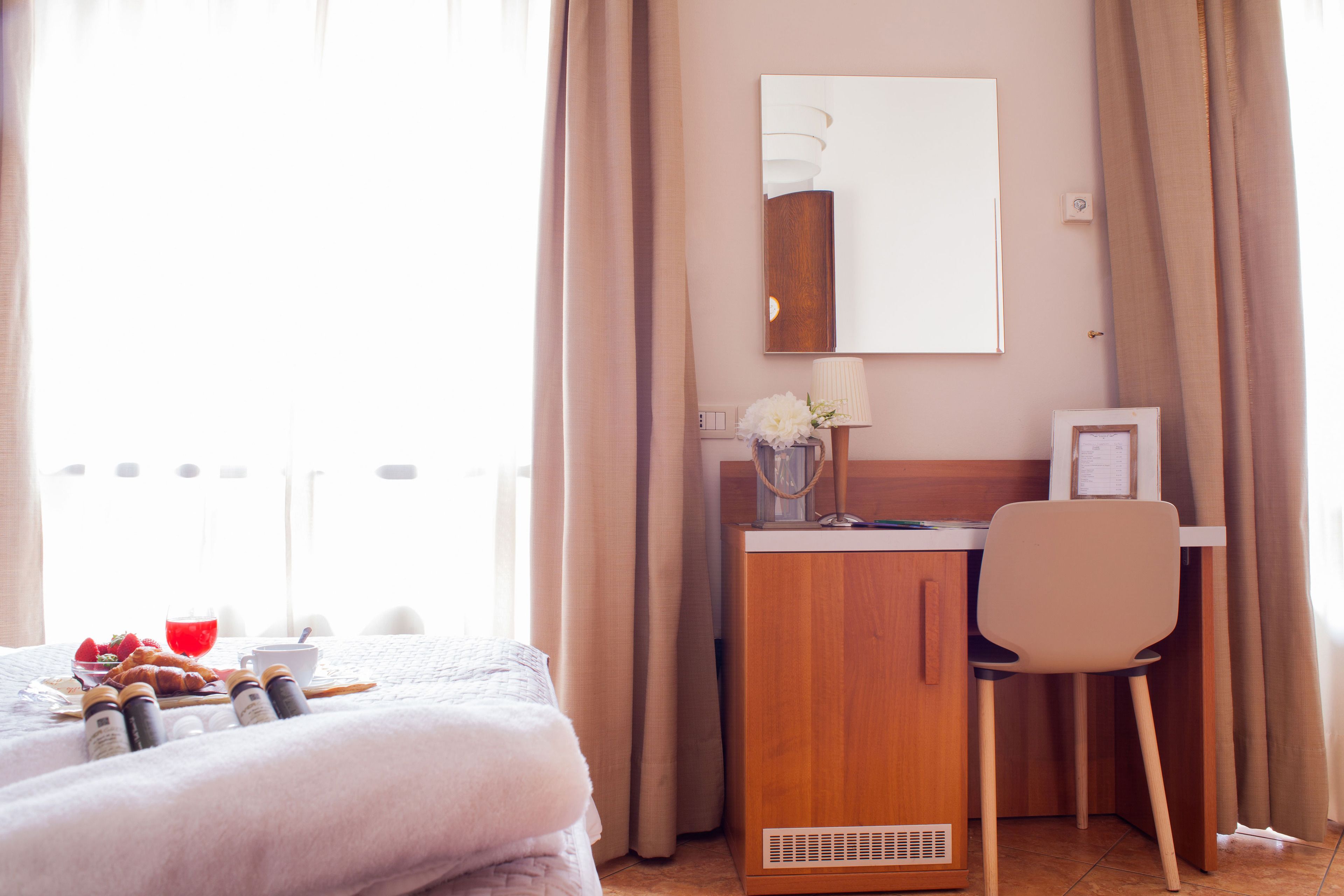 A hotel room with a bed, a desk with a chair, a mirror, and a window with curtains.