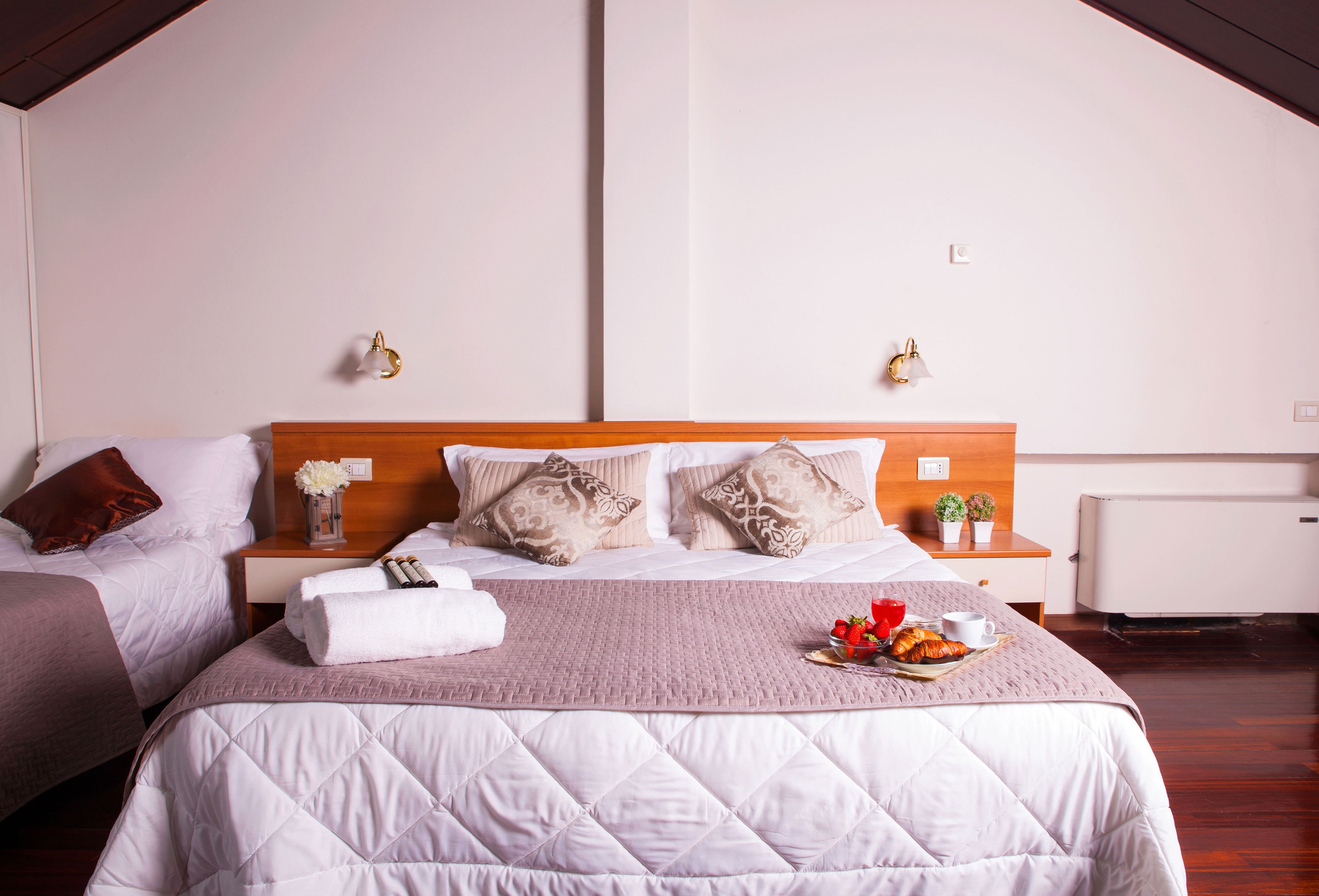 Quarto de hotel com duas camas, cabeceira de madeira, mesa de cabeceira com um vaso de flores e bandeja com morangos e uma chávena.