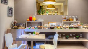 A breakfast buffet with various food items and beverages, a seating area with white chairs, and a mirror reflecting the room. - Hotel Ares (Milan)