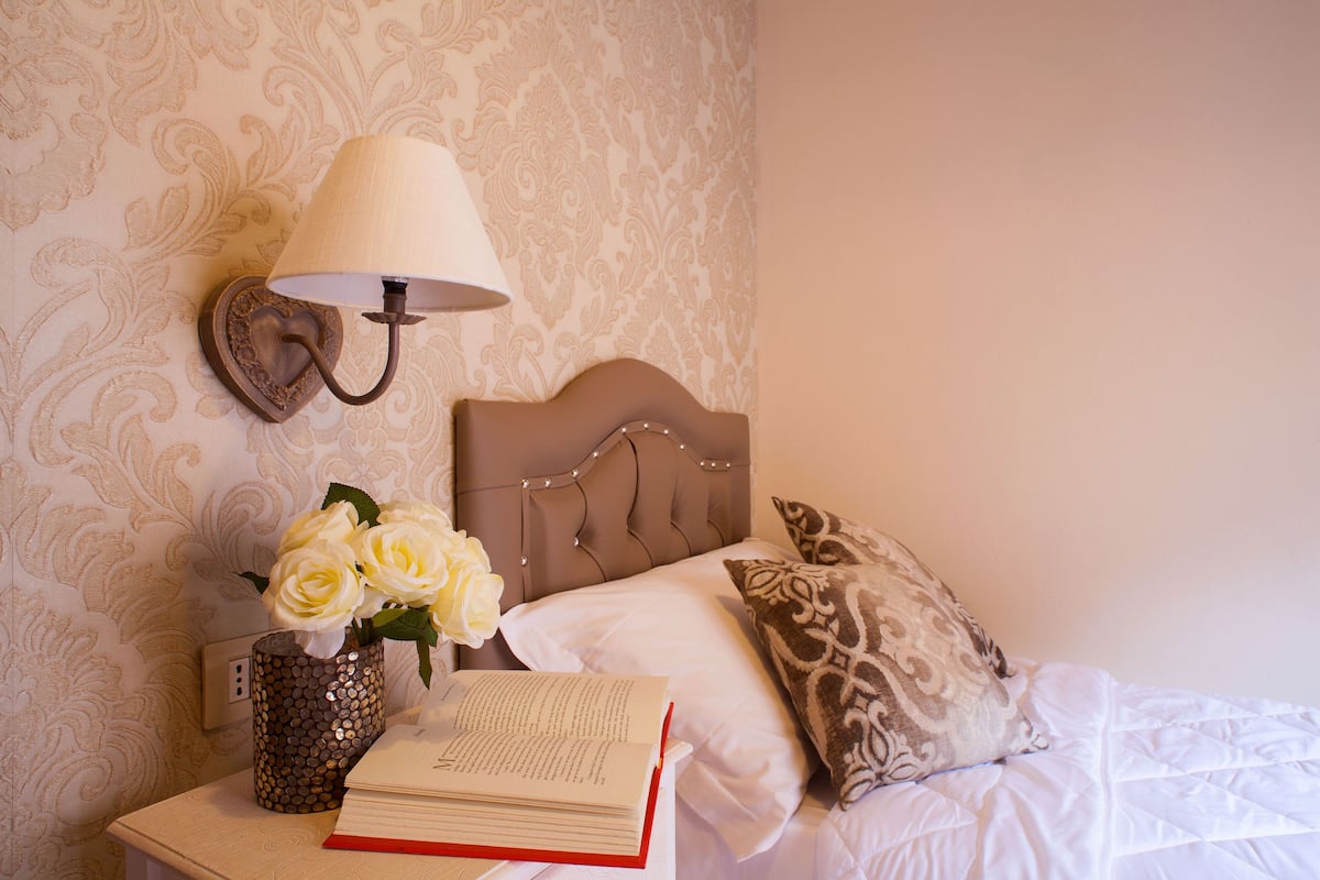 A neatly made bed with a patterned pillow, a bedside lamp, a vase with flowers, and an open book on a bedside table.