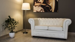 A white tufted sofa, a floor lamp, and a potted plant in a room with a dark wall and a framed artwork. - Hotel Ares (Milan)