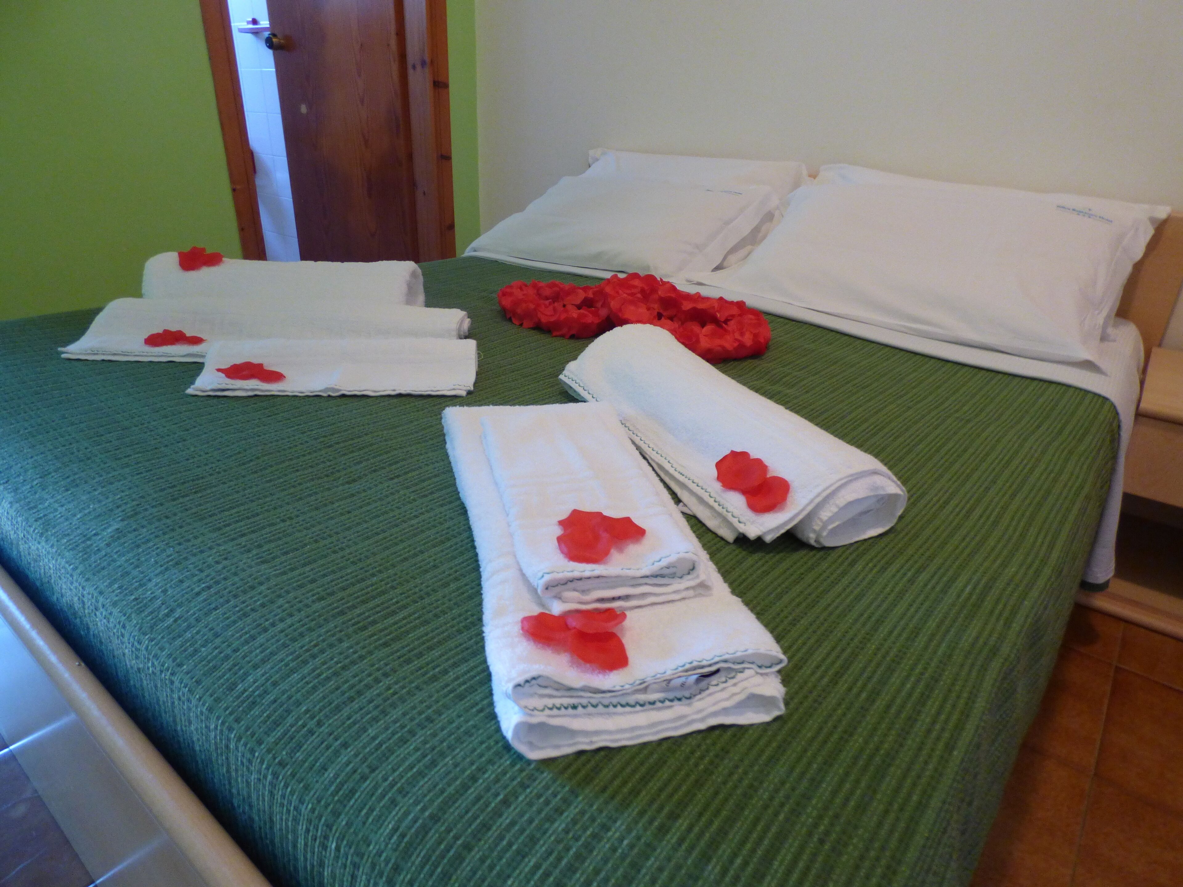 A neatly made bed with green bedding, white pillows, and rolled towels with red flowers.