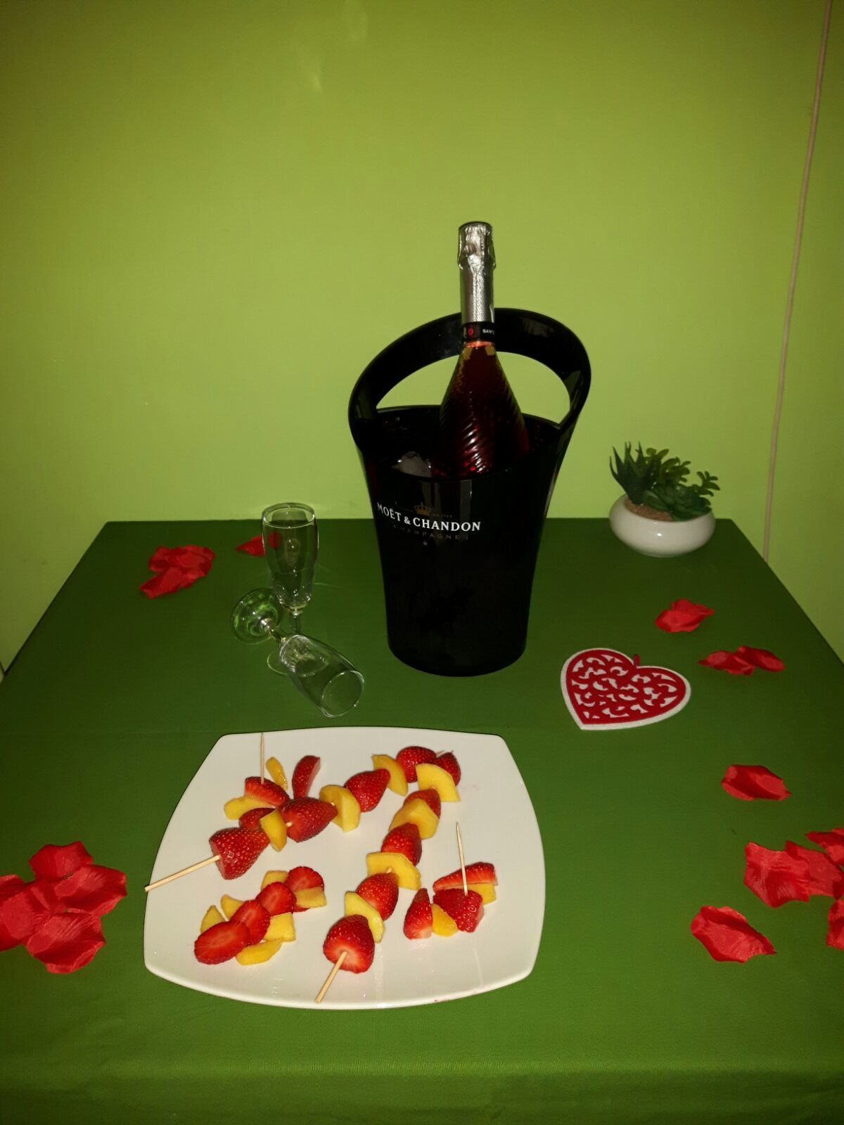 A table with a bottle of Moët & Chandon champagne, a glass, a small potted plant, a plate of fruit skewers, and a heart-shaped decoration.