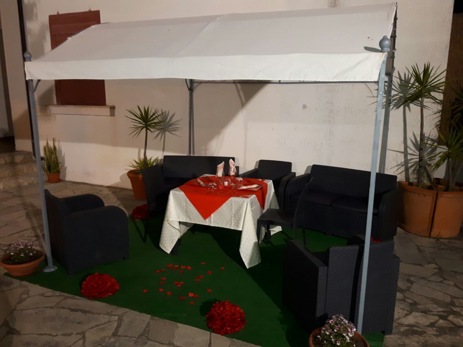 Ein Essbereich im Freien mit einem Tisch für zwei Personen, umgeben von schwarzen Stühlen, unter einer weißen Markise. Übertöpfe mit Pflanzen und ein grüner Teppich mit roten Blüten liegen auf dem Boden.