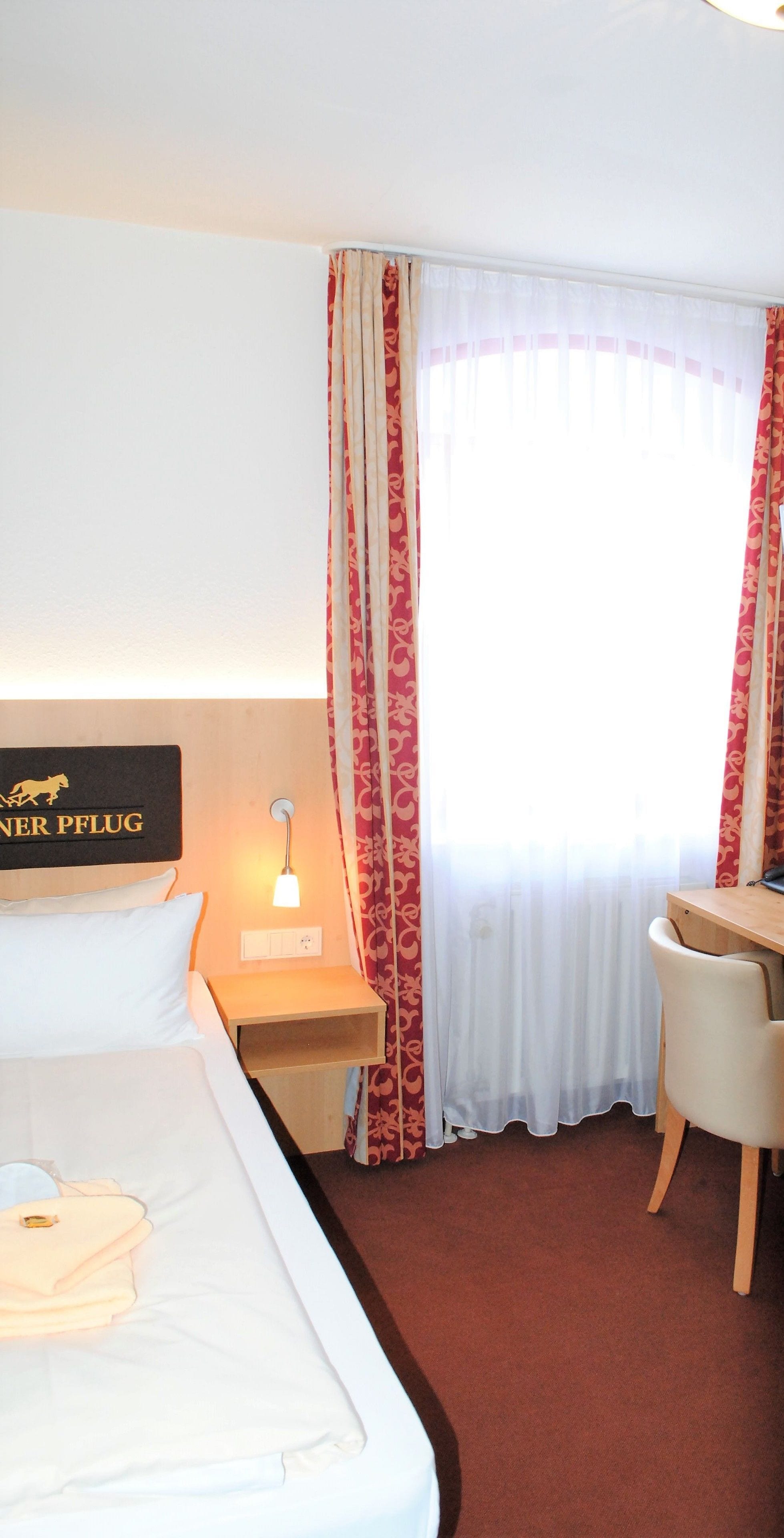 A hotel room with a bed, a desk, a chair, a nightstand with a lamp, and a window with curtains.
