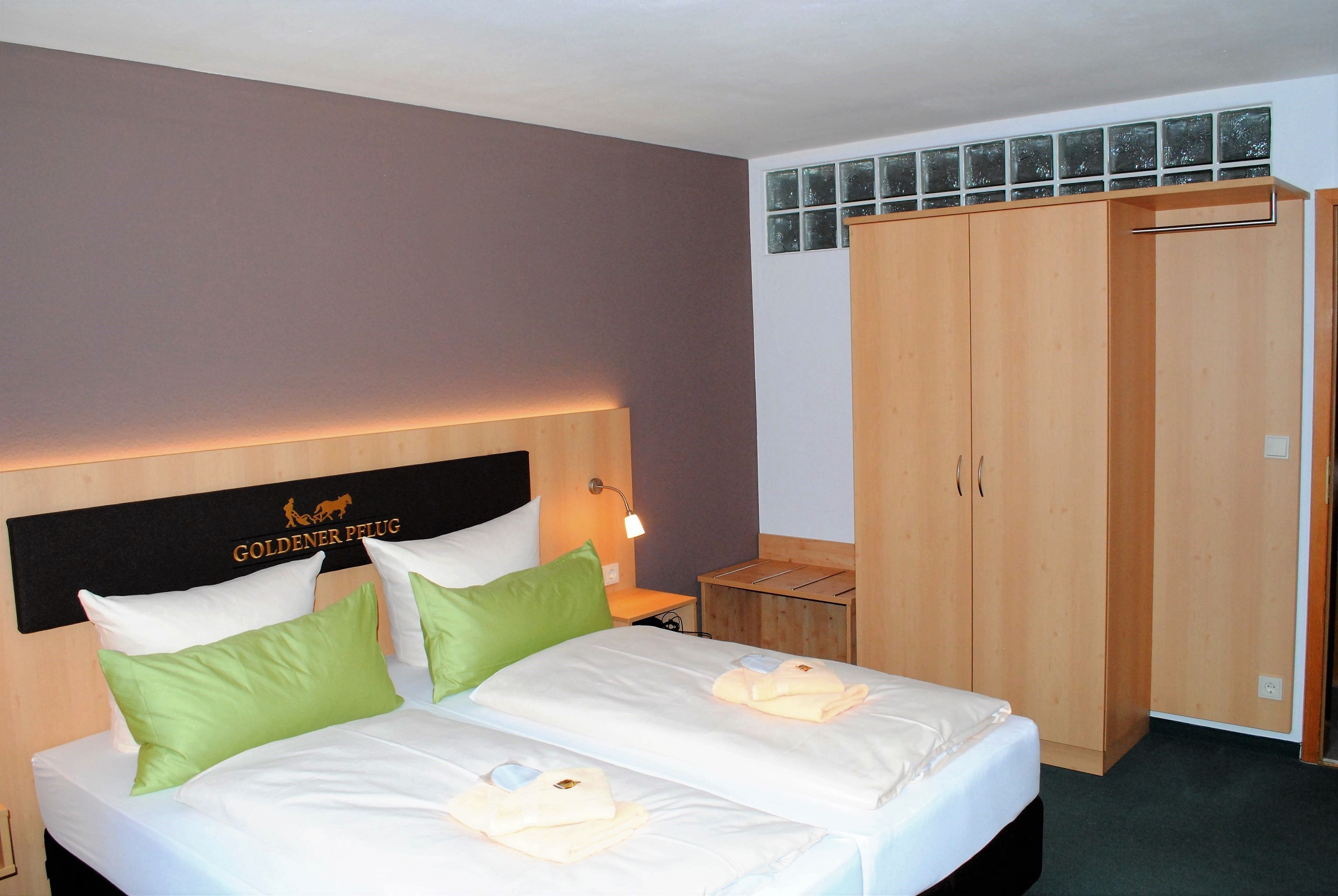 Une chambre d’hôtel avec deux lits, une tête de lit ornée d’un logo, une armoire et une table de chevet avec une lampe.