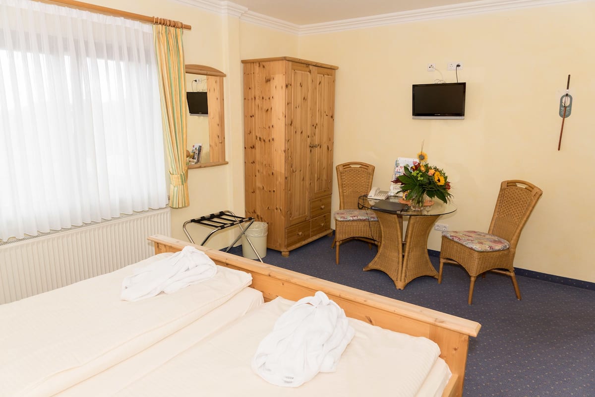 A hotel room with two beds, a wooden wardrobe, a television, a small table with a vase of flowers, and wicker chairs.