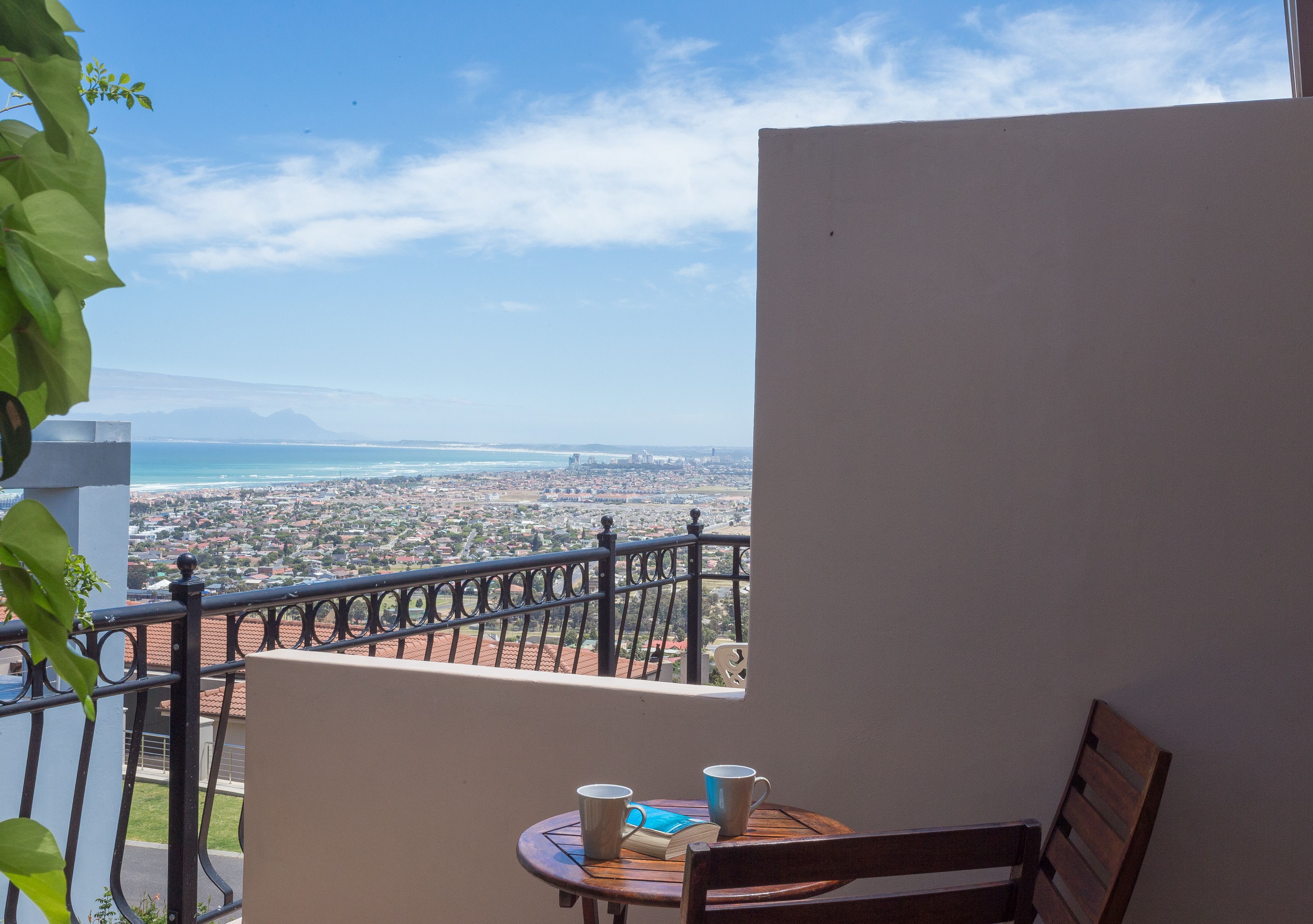 Ein Balkon mit Blick auf eine Küstenstadt, ein Tisch mit zwei Tassen und einem Buch sowie ein Holzstuhl.