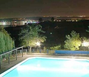 Una piscina nocturna con árboles circundantes y luces de la ciudad a lo lejos.