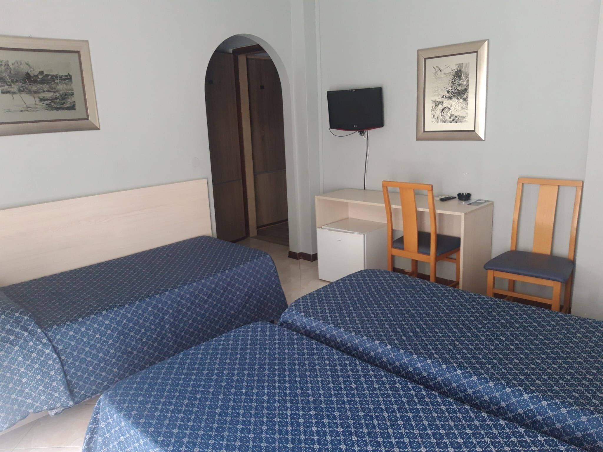 Habitación de hotel con dos camas individuales, un escritorio, una silla, un televisor y un arco de entrada.