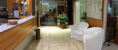 Un vestíbulo de hotel con mostrador de recepción, una máquina de café, una planta en maceta y dos sillones blancos.