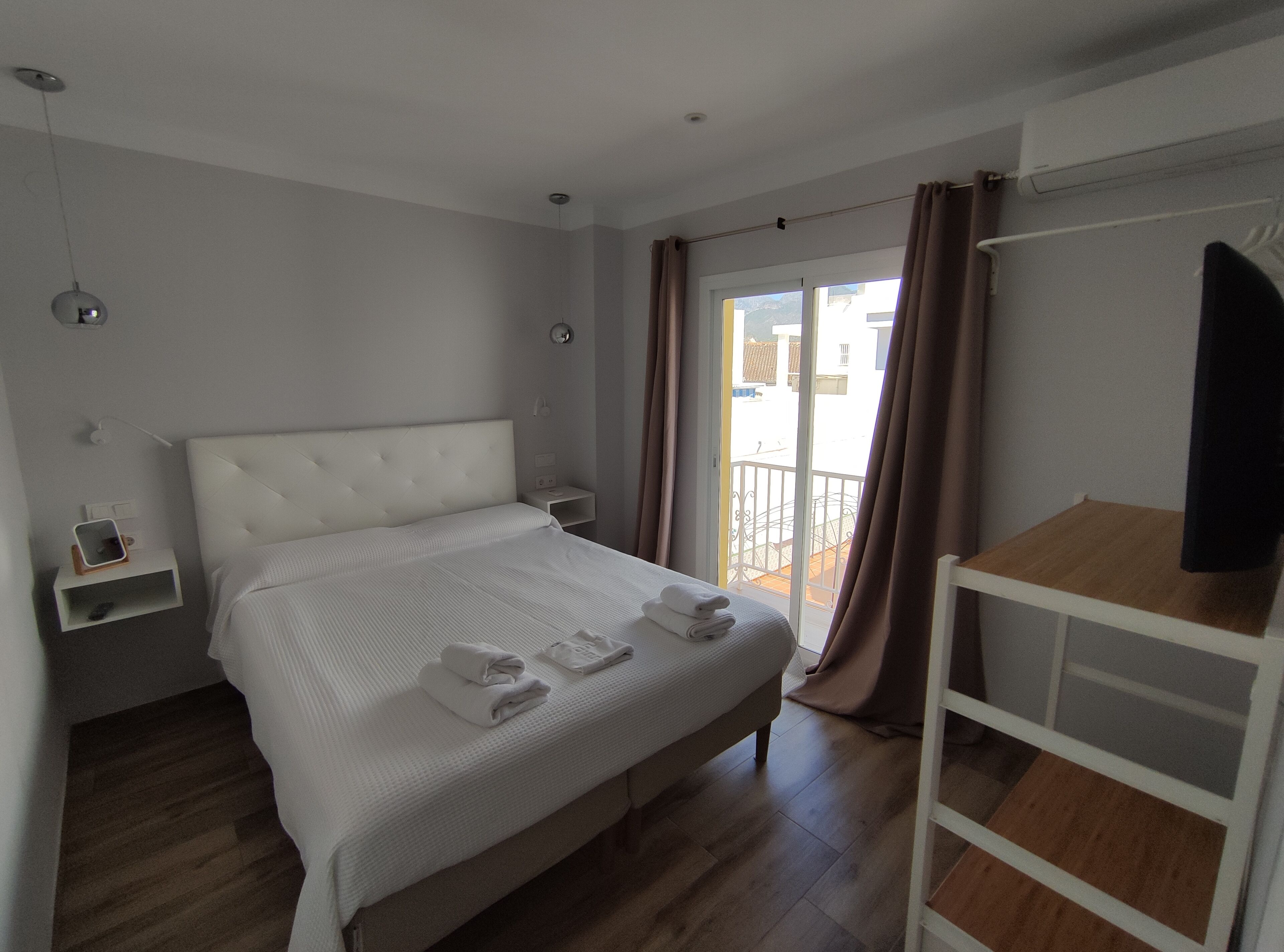 Une chambre d’hôtel moderne avec un grand lit, une table de chevet, une petite étagère et une vue sur le balcon extérieur.