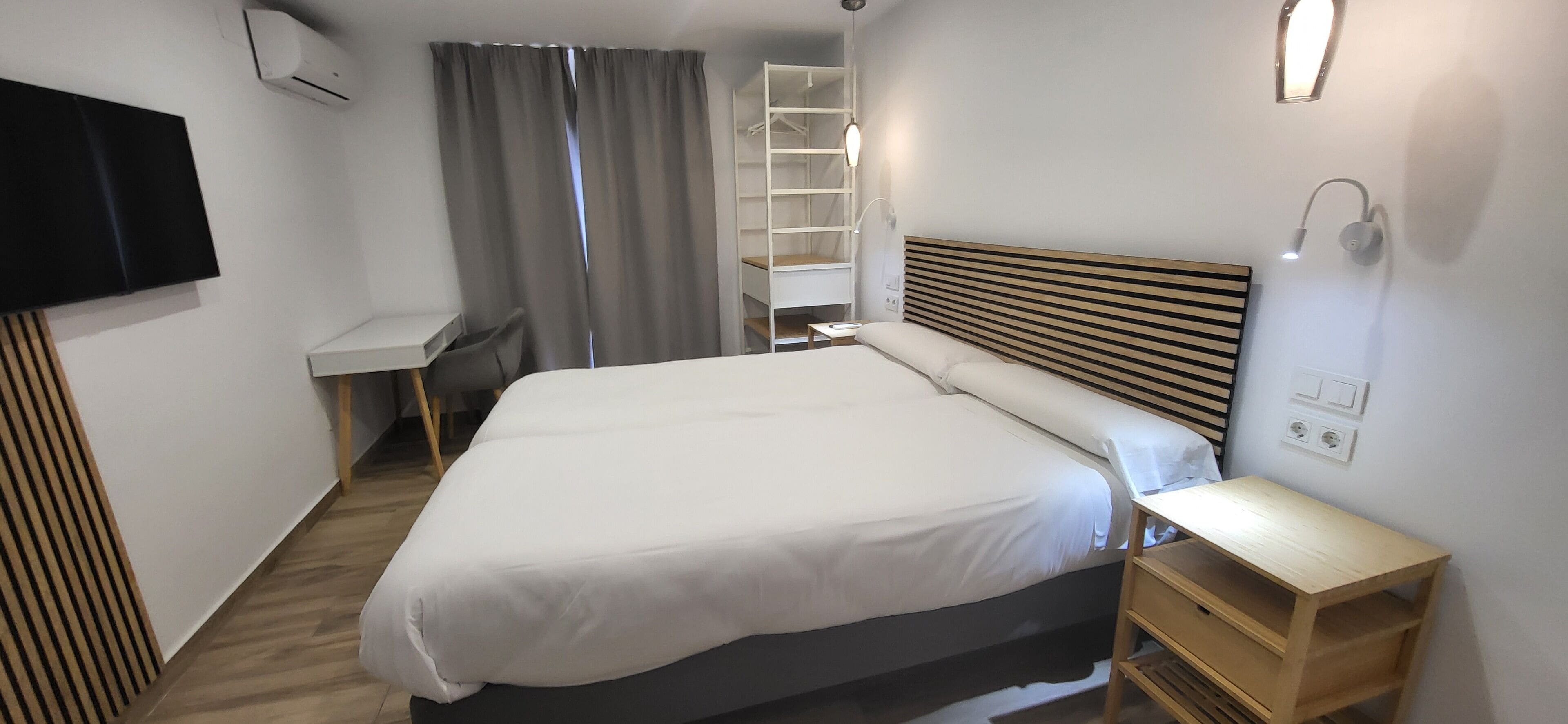 A modern hotel room with a large bed, a bedside table, a TV mounted on the wall, and a built-in shelving unit.